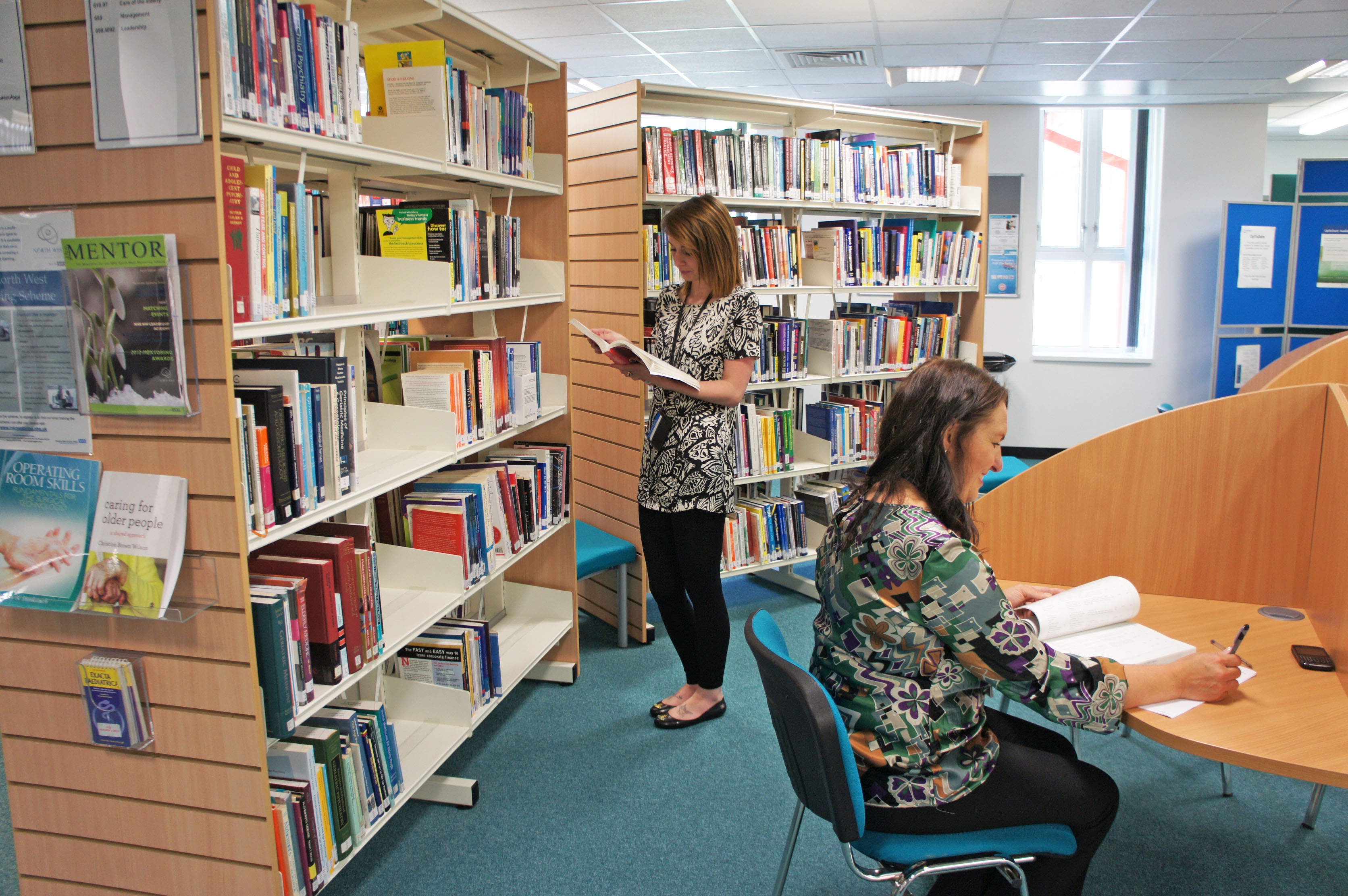 Two women are in a library. One is sat down with pen in hand studying. The other is stood in front of library book shelf reading a book.